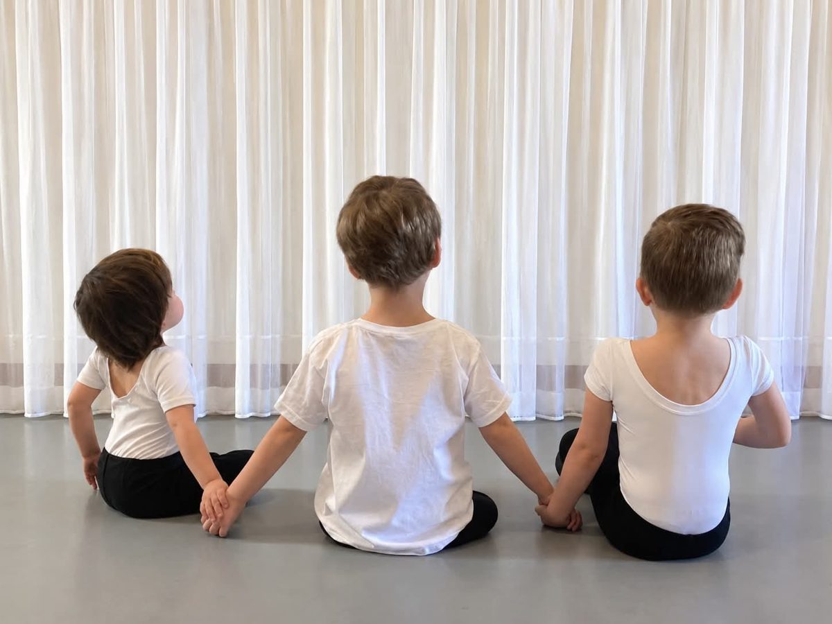 Three young boys seen from behind in white T-shirts and black shorts, sitting on the studio floor holding hands
