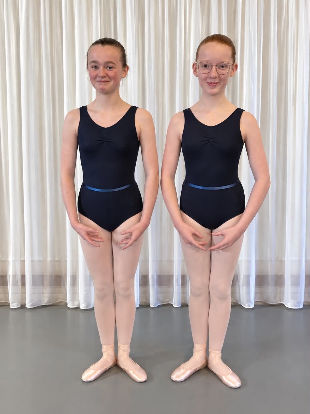 Two upper school ballet students in navy leotards with blue elastic belts, pink tights and pink ballet shoes