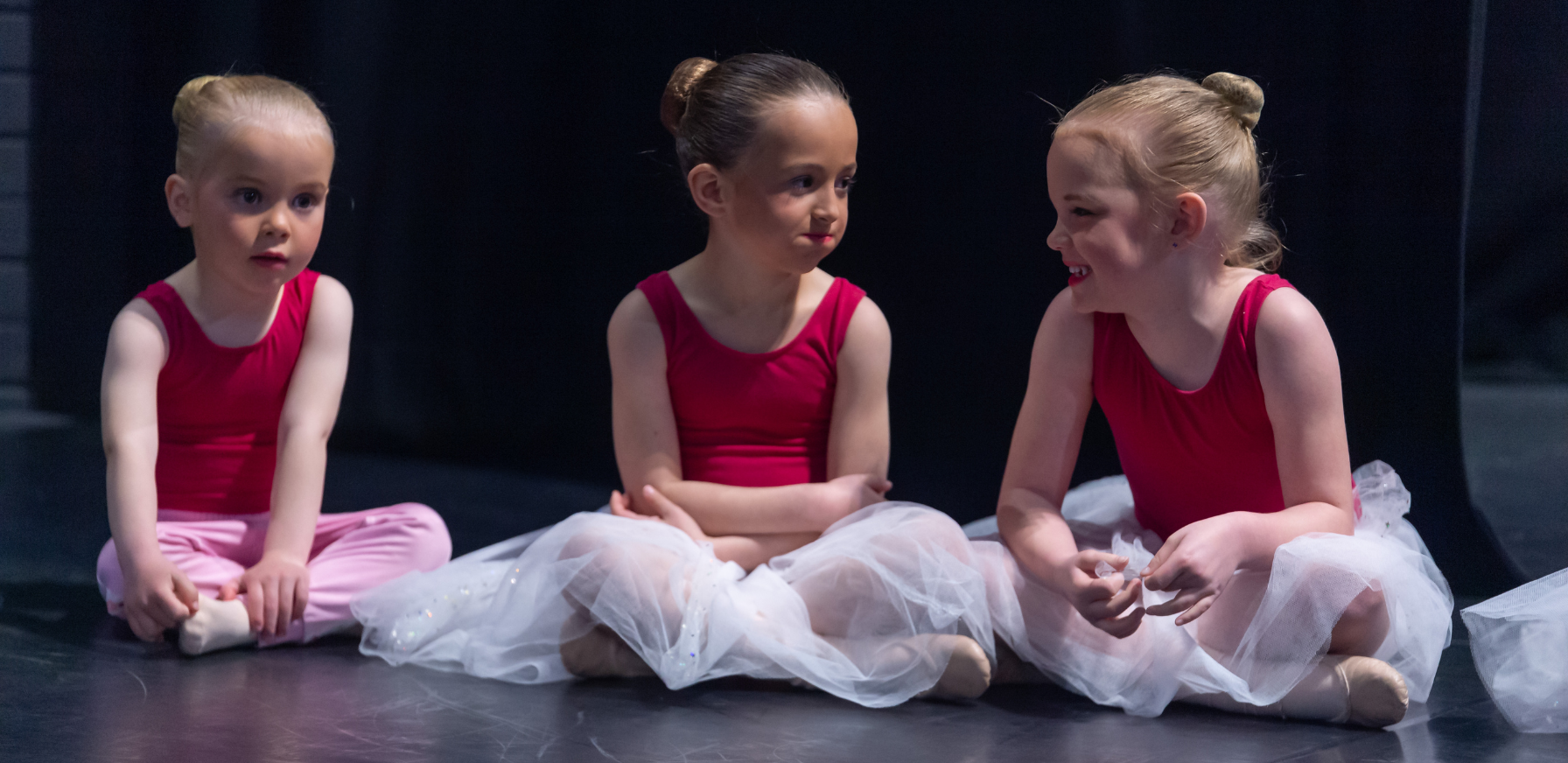 Three young ESSOPA ballet students sitting together on stage in red leotards and white tutus, laughing