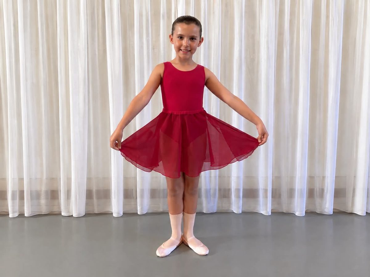 A young ballet student in a red leotard with attached chiffon skirt, pink socks and pink ballet shoes